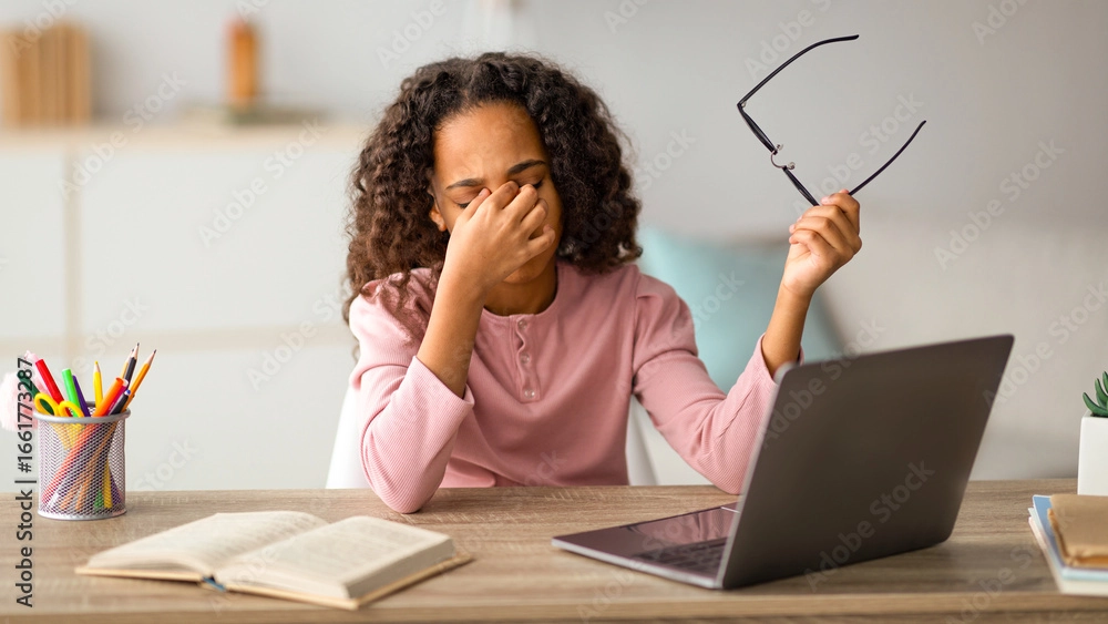 A young black woman sits at a desk in front of her laptop, rubbing her eyes and holding her glasses to one side.