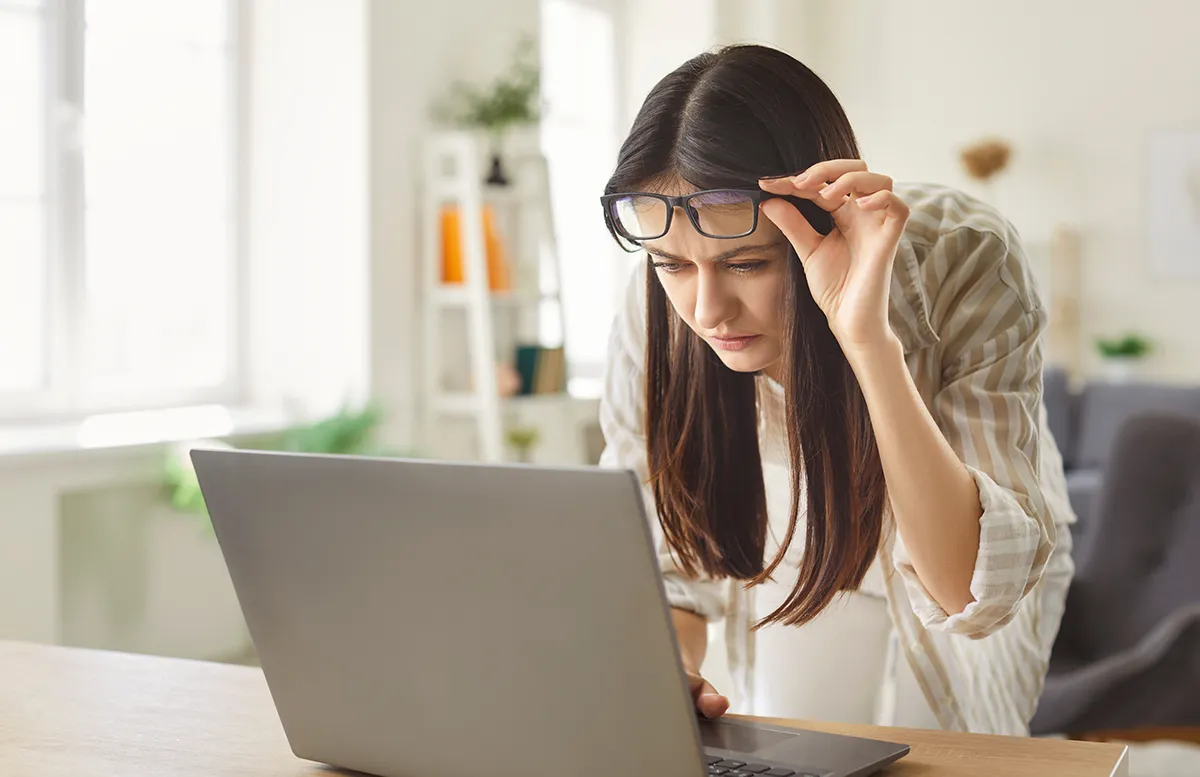 A young woman sits at a desk in front of her laptop, lifting up her glasses.
